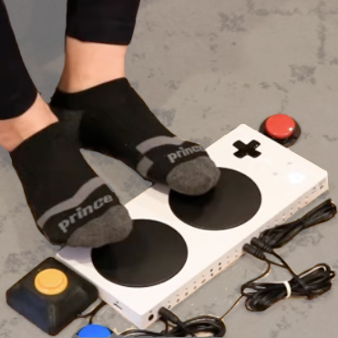 A person's feet interacting with the buttons on the Xbox Adaptive Controller, with several button switches surrounding it.